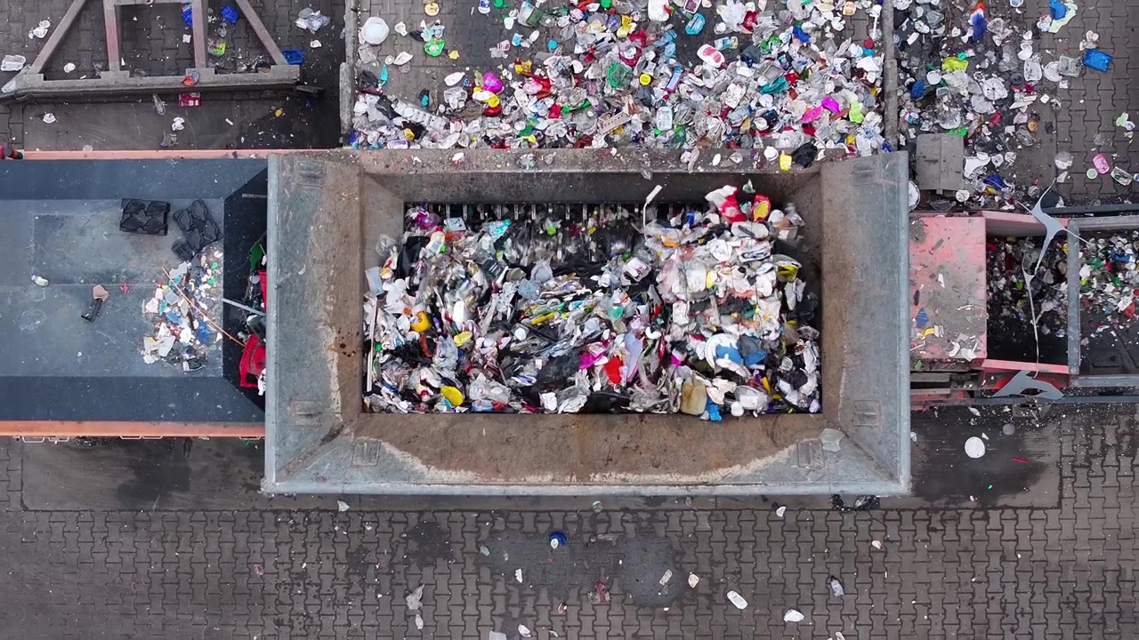I rifiuti di plastica entrano in un grande contenitore presso un impianto di riciclaggio. I lavoratori smistano vari tipi di rifiuti per il riciclo. Le operazioni mirano a un efficiente recupero dei materiali e alla riduzione dei rifiuti