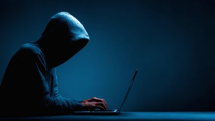 Una figura misteriosa in felpa con cappuccio dedita a un complesso lavoro al computer sotto una luce fioca, che incarna l'essenza dell'intrigo cibernetico e del mistero.