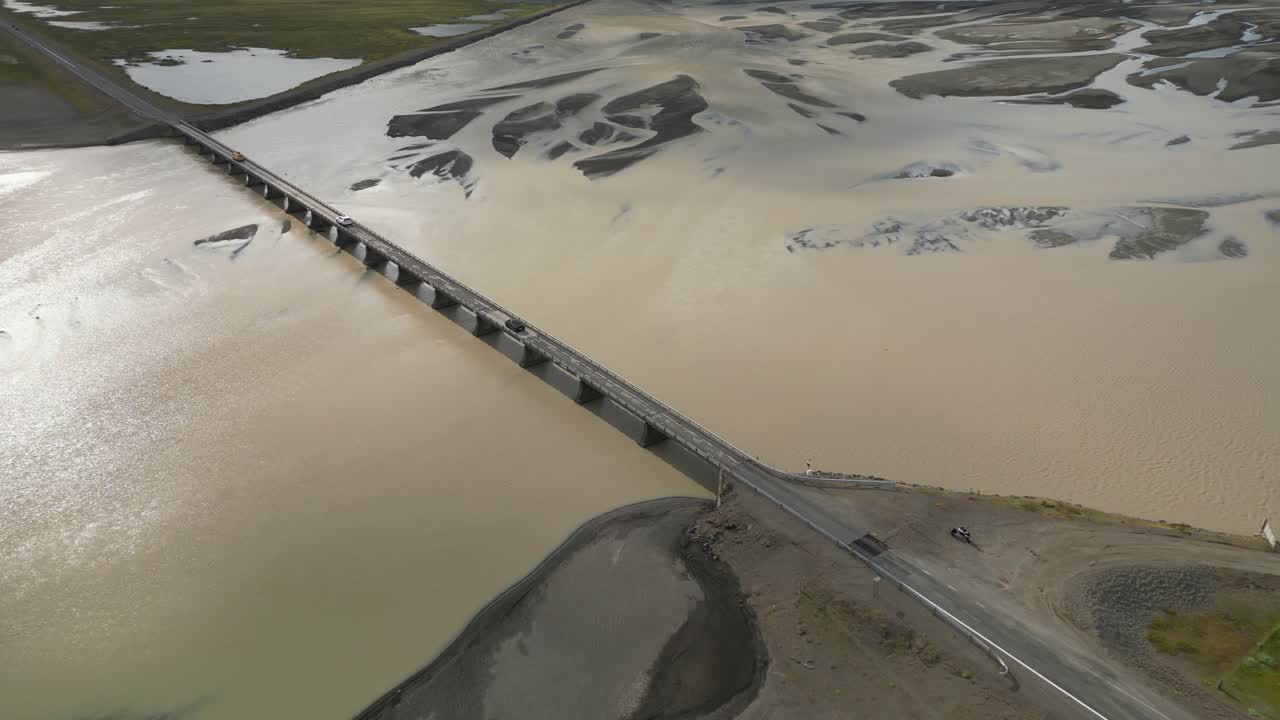 바다 쪽으로 천천히 흐르는 어두운 빙하 강을 가로질러 걸려있는 다리에서 운전하는 자동차의 공중 전망