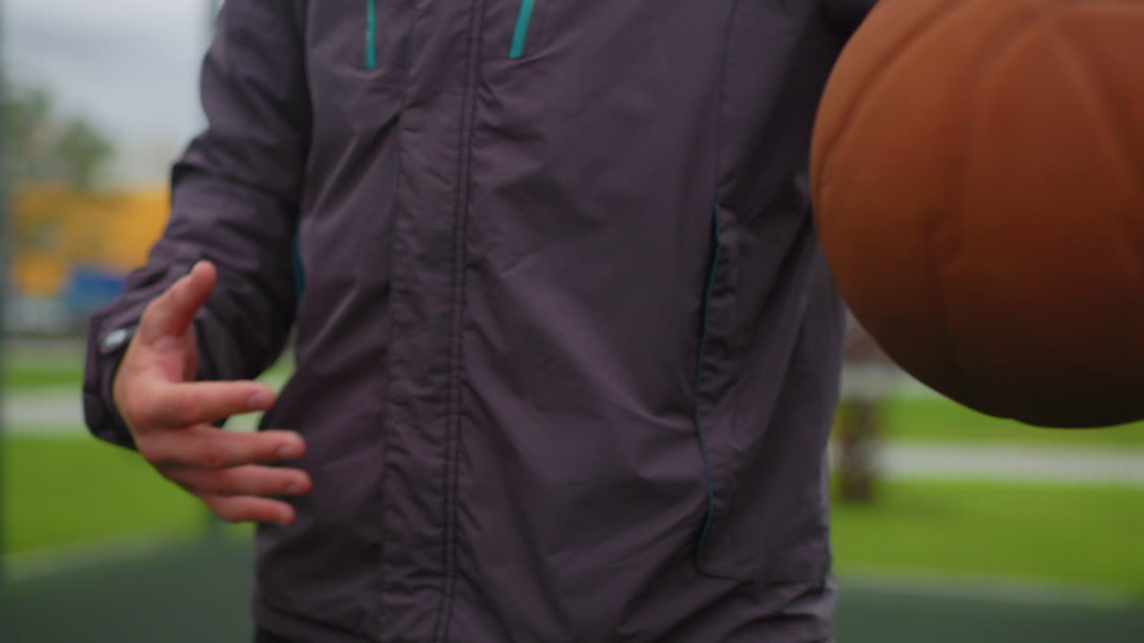 young player dribbling basketball on court, gray jacket and windbreaker, closeup hand control and finger grip, overcast sky and metal fence, distant industrial buildings and parking lot, casual