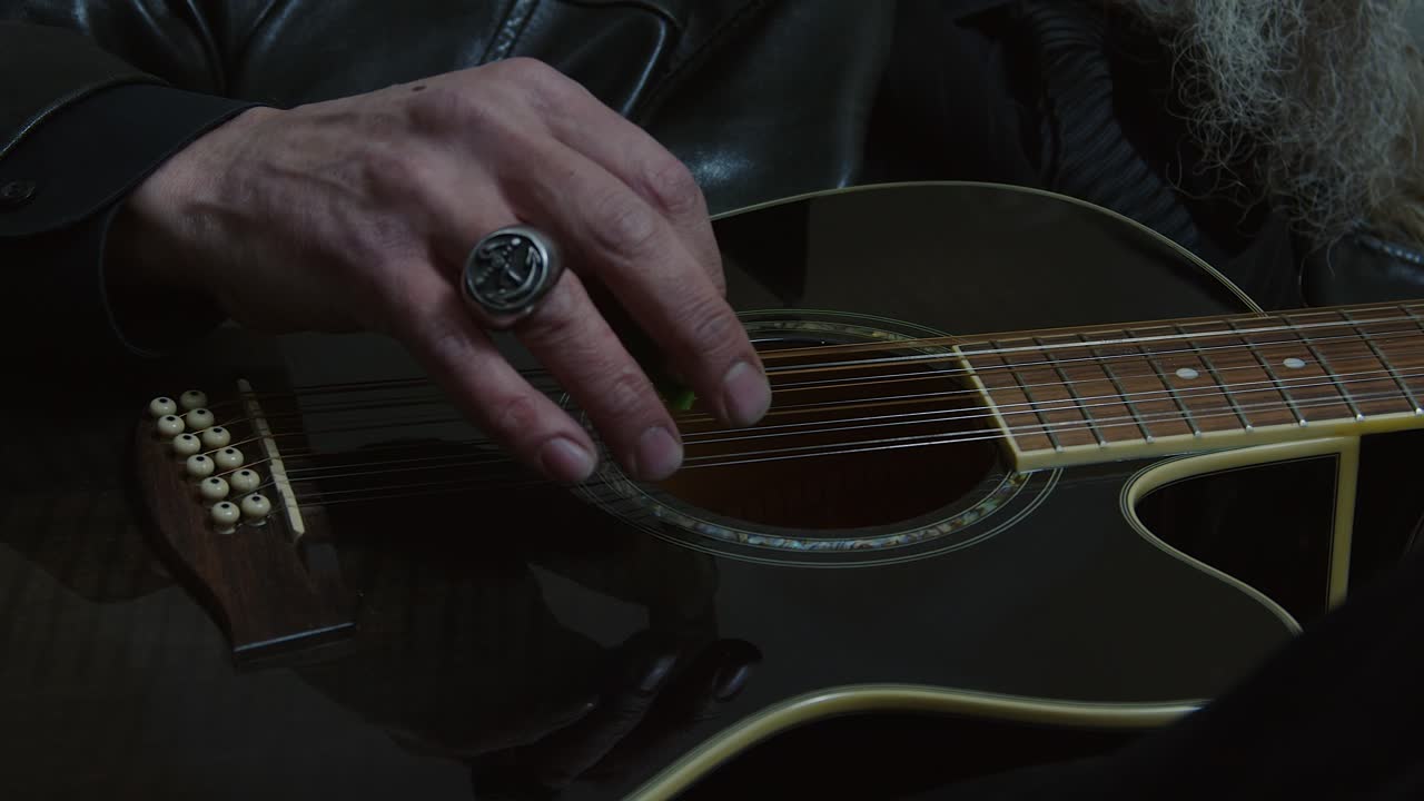 Closeup of hand: Bearded grey haired man plays black 12-string guitar