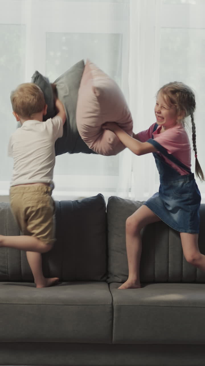 Brother and sister fight with pillows standing on soft couch against bright window. Cheerful siblings have fun at home after preschool