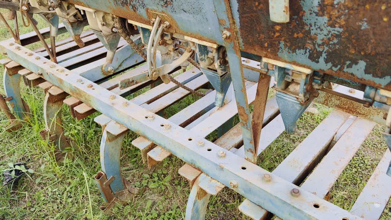 A rotating camera movement around an old rusty blue seed drill machine, revealing its corroded metal surface, gears, and levers — a glimpse of aged farming technology standing still in time