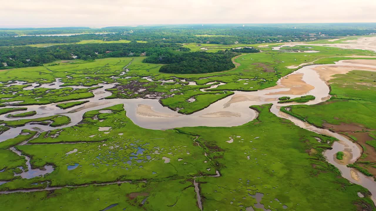Aerial perspective reveals marshland with tidal flats, sandbars, and winding creeks weaving across the wetlands, highlighted by reflective pools glistening in soft natural light