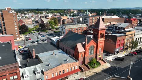 Aerial-view-of-an-urban-landscape-with-historic-red-bricked-church,-diverse-buildings,-parking-lots,-and-streets-with-parked-cars-under-a-clear-sky