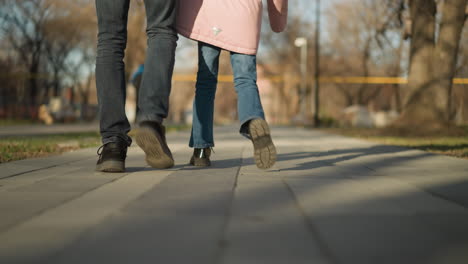 focused view of a little girl and an adult man's legs walking together on a park pathway, both wearing blue jeans and black shoes in daylight