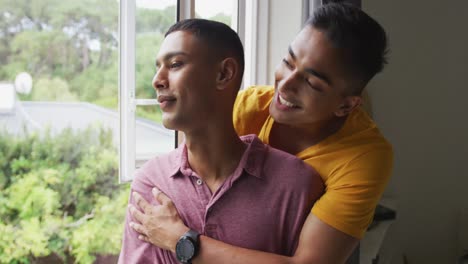 Happy-mixed-race-gay-male-couple-standing-looking-out-of-window-and-embracing