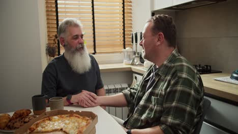 Happy-couple-of-LGBT-men-talking-and-holding-hands-while-sitting-in-the-kitchen-during-their-lunch.-An-elderly-man-with-gray-hair-and-a-lush-beard-in-a-gray-T-shirt-holds-hands-with-his-brunette-boyfriend-in-a-checkered-green-shirt-while-sitting-in-the-kitchen-at-the-table