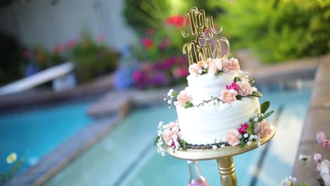 shot of a beautifully decorated floral wedding cake sitting on a pedestal right next to a pool during a summer wedding reception