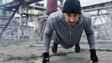 guapo boxeador masculino con gorro haciendo flexiones antes de entrenar boxeo al aire libre en la fábrica abandonada en una mañana nublada