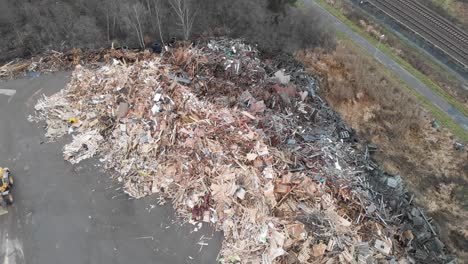 Weitwinkelflug-In-Geringer-Höhe-Auf-Riesige-Deponiehaufen-Voller-Tonnen-Von-Holz--Und-Sperrmüll-Aus-Europaletten,-Während-Traktor-Sägemehl-Schaufelt