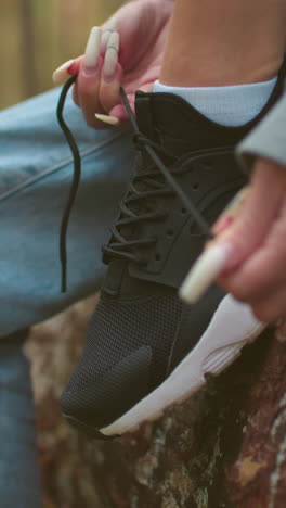 primer plano de una mujer con largas uñas blancas atando cordones de zapatillas en zapatillas negras, con suéter gris y vaqueros azules claros, sentada en un árbol caído durante una caminata en el bosque