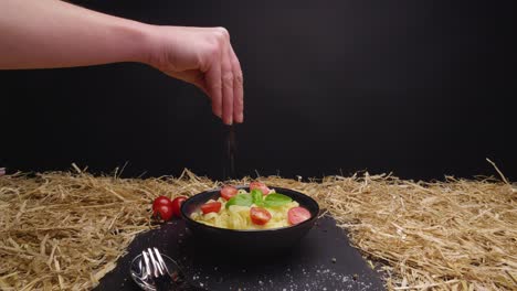 dolly shot of a bowl on a slate with delicious fresh pasta seasoned with basil and herbs with a hand seasoning the food against black background