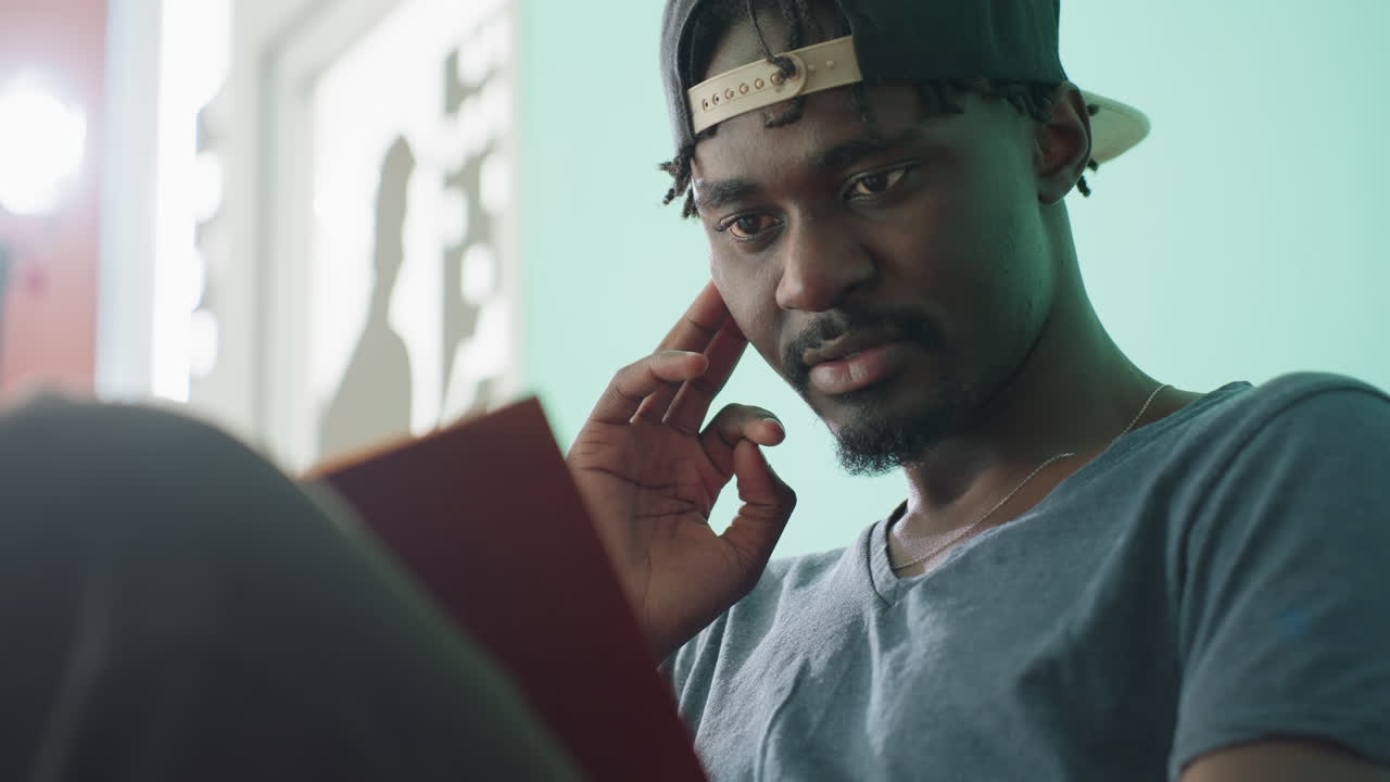 Close up of focused freelancer wearing cap reading book with thoughtful expression while touching nose as soft light from window creates gentle glow and shadow silhouette on wall in calm indoor space