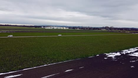 Drohnenaufnahme-Von-Braunen-Und-Grünen-Feldern-Im-Winter-Mit-Wenig-Schnee-Mit-Schneller-Unterer-Kreisbewegung-Nach-Rechts-Und-Fahrenden-Autos-Auf-Der-Straße