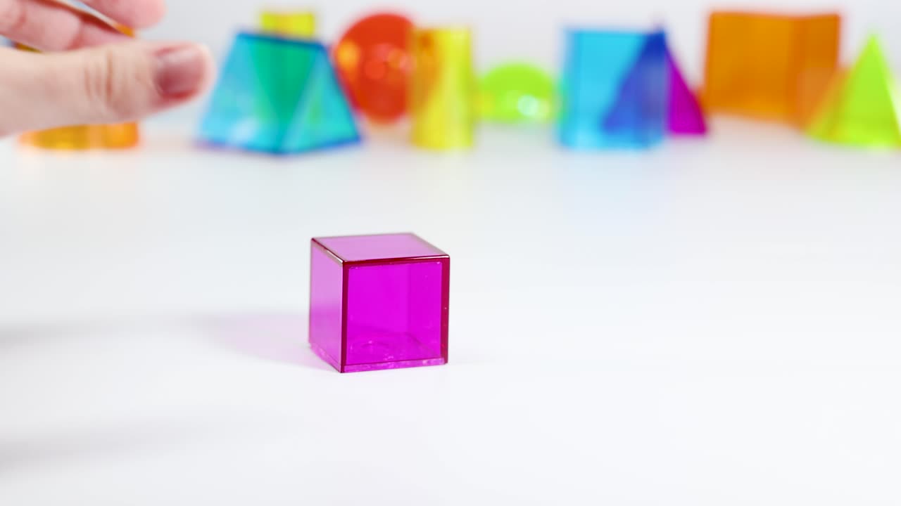 A hand manipulates a pink cube among vibrant 3D shapes on a white surface, highlighting geometry and color