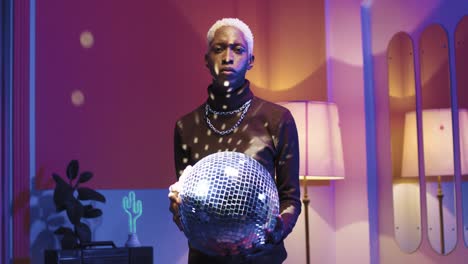 young stylish man holding disco ball in his hands and looking at camera in a retro party at home