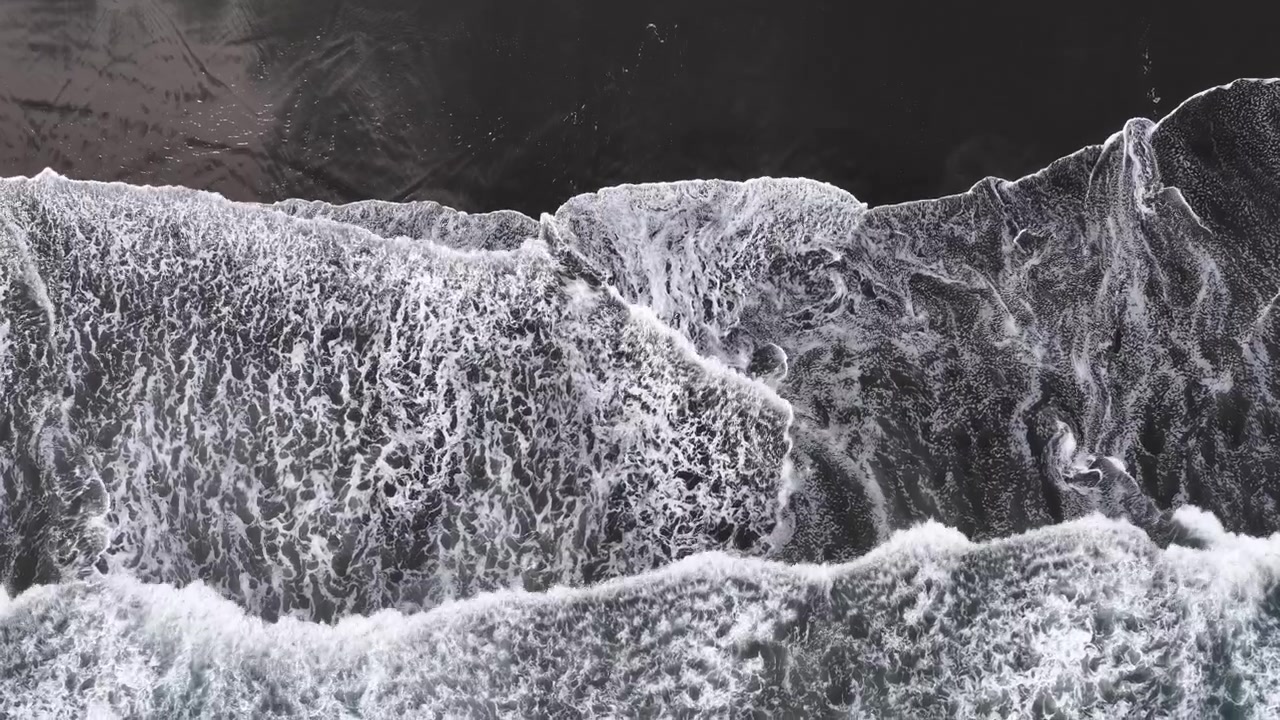 Drone footage captures waves crashing onto a dark volcanic sand beach on New Zealand's North Island.