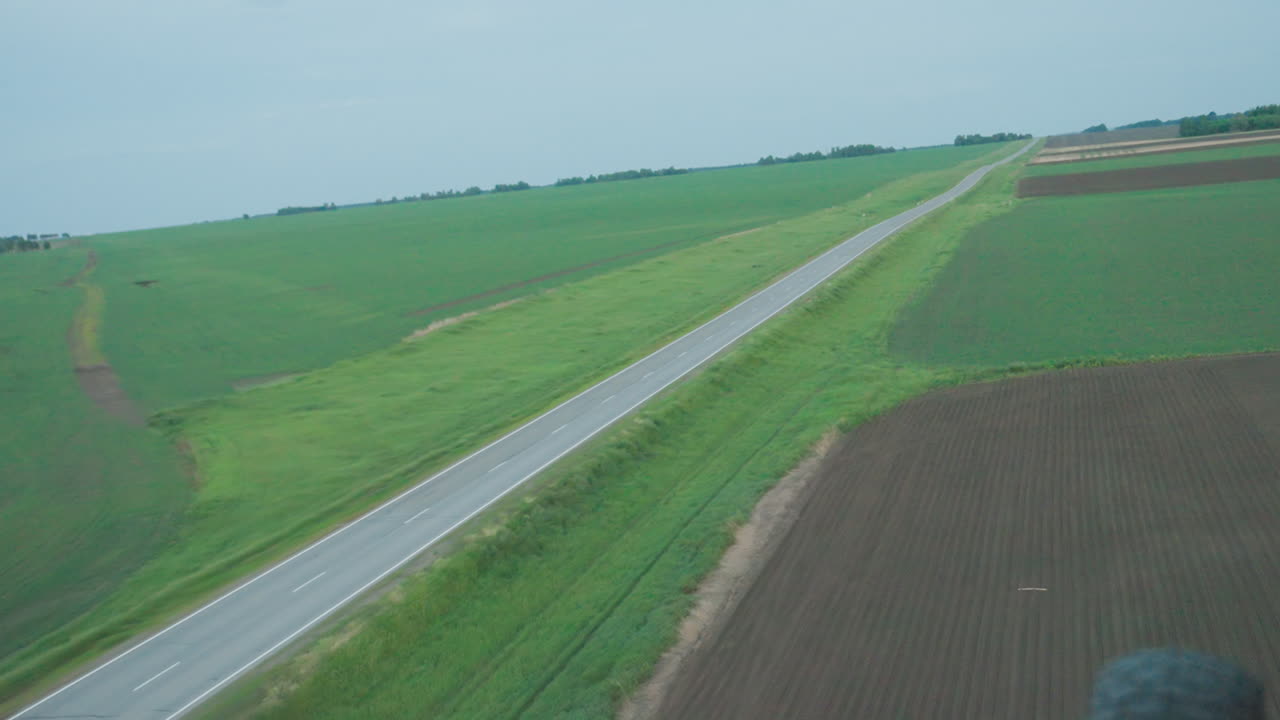 overhead drone view of straight rural road cutting through lush green farmland under soft daylight, white dashed markings drawing visual path across emerald fields toward distant horizon