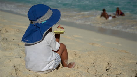 Unrecognizable-mexican-latin-boy-with-a-hat-and-a-whit-t-shirt-sitting-on-the-sand-playing-with-a-plastic-shovel-watching-the-ocean