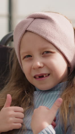 toothless girl with paralysis turns to camera showing thumbs up gesture on blurred background. preschooler kid sits in wheelchair on street closeup