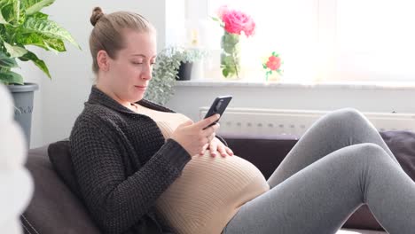 Schwangere-Weiße-Frau-Sitzt-Auf-Der-Couch,-Tippt-Auf-Das-Telefon,-Streichelt-Ihren-Bauch-Und-Lächelt-In-Zeitlupe