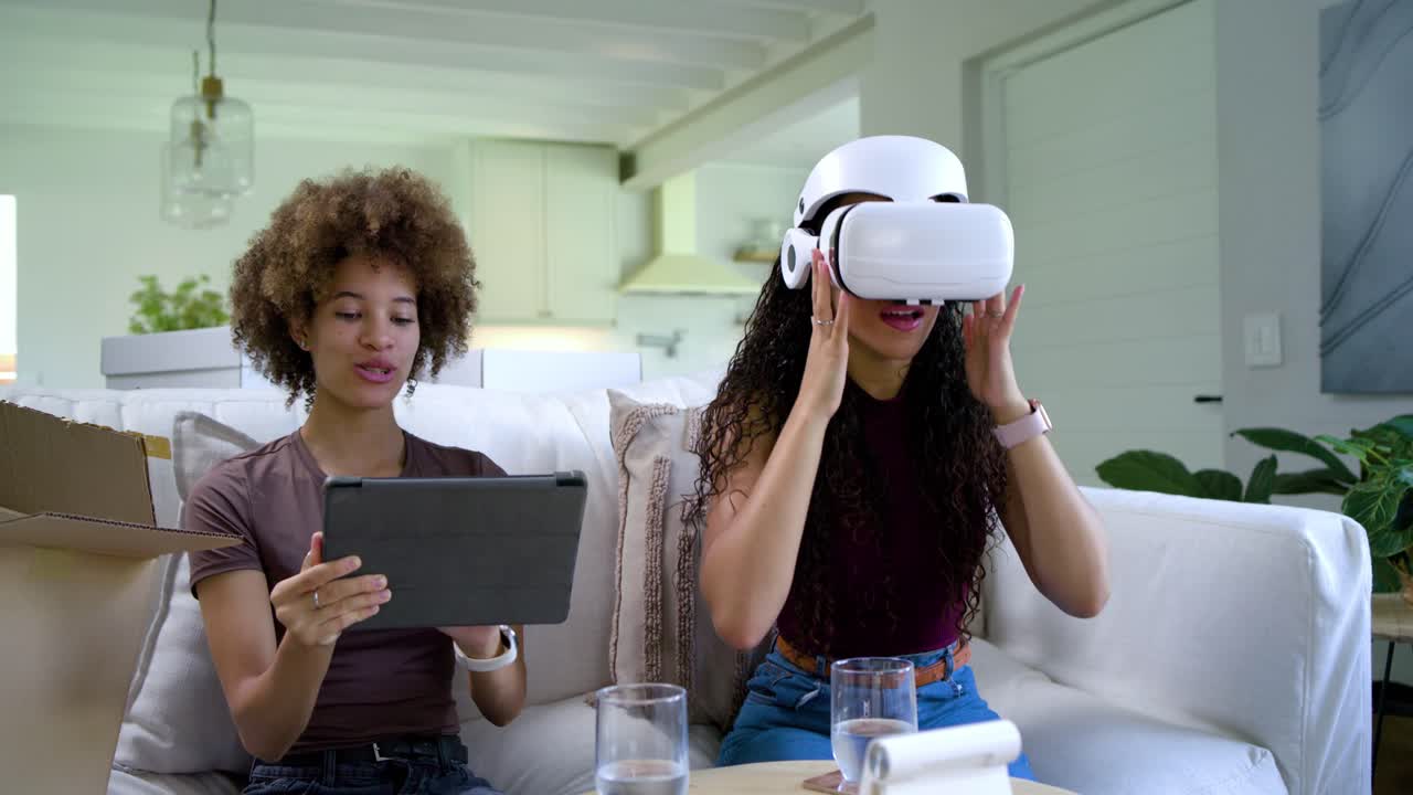 African American female friends unboxing on sofa, right adjusting VR headset, left tapping tablet