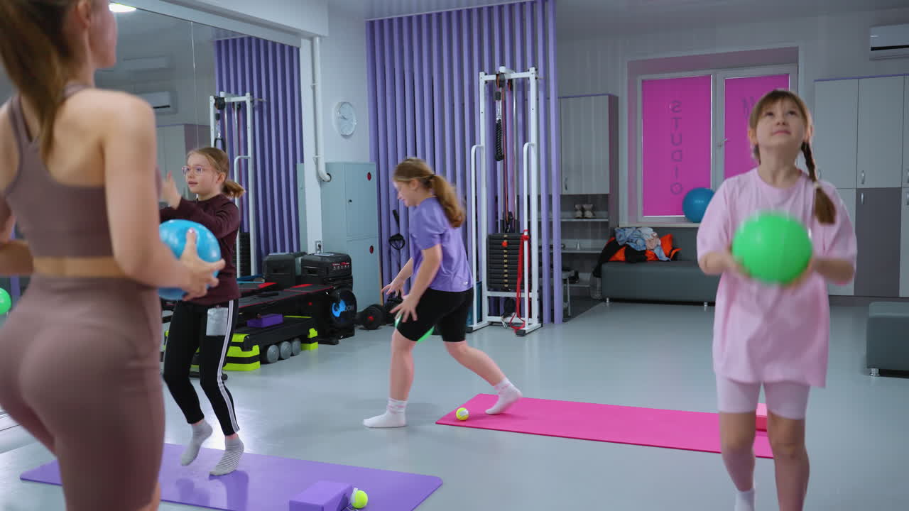 instructor en traje marrón malabarismo, lanzando globo con un niño pequeño tratando de imitar la acción, globo cayendo de la mano del niño en un gimnasio moderno con otros niños y equipos de gimnasio en el fondo
