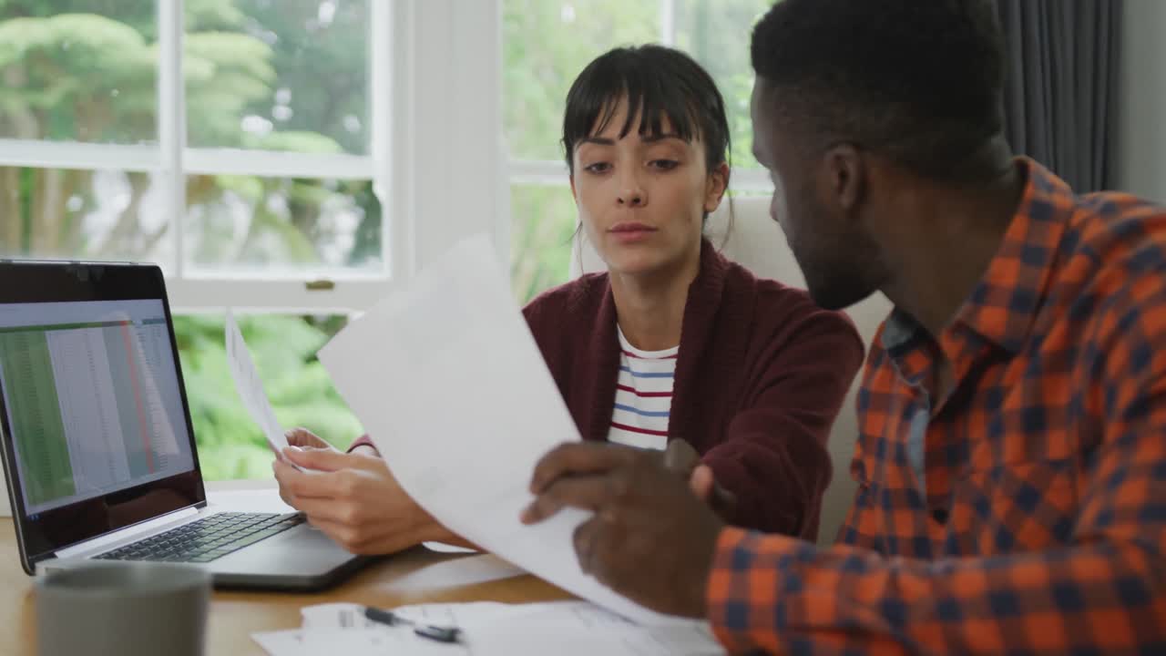 Diverse couple sitting at table talking and working with laptop