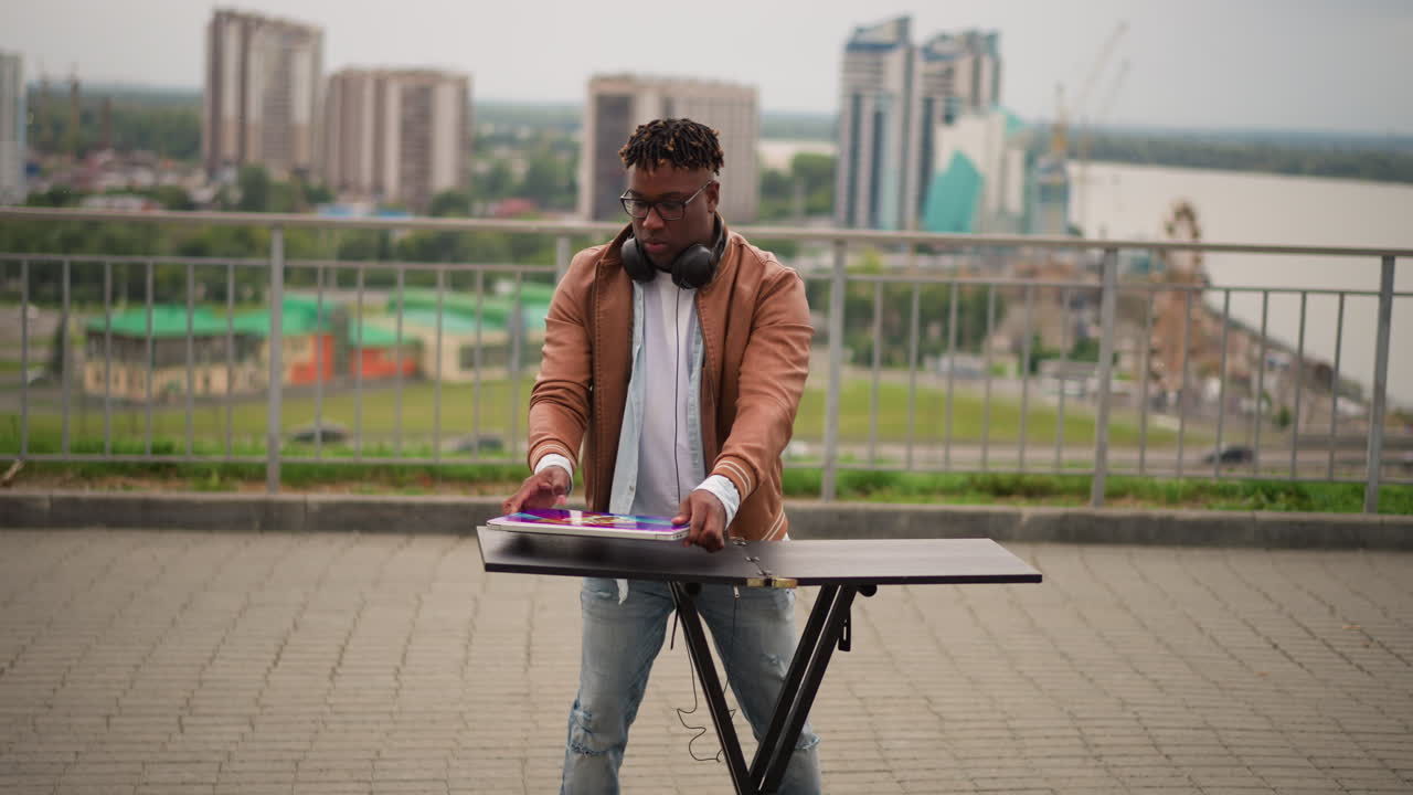 Black discjockey setting up rooftop table overlooking city skyline. Solo street musician retrieves gear from red bag, assembles mixer and controller on compact stand, adjusts headphones and tests