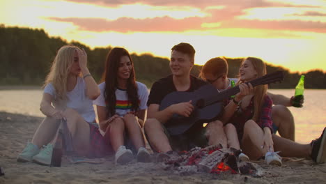 The-company-of-people-plays-the-guitar-and-sings-songs-around-bonfire-on-the-beach.-This-is-perfect-summer-evening-with-the-best-friends-and-beer.