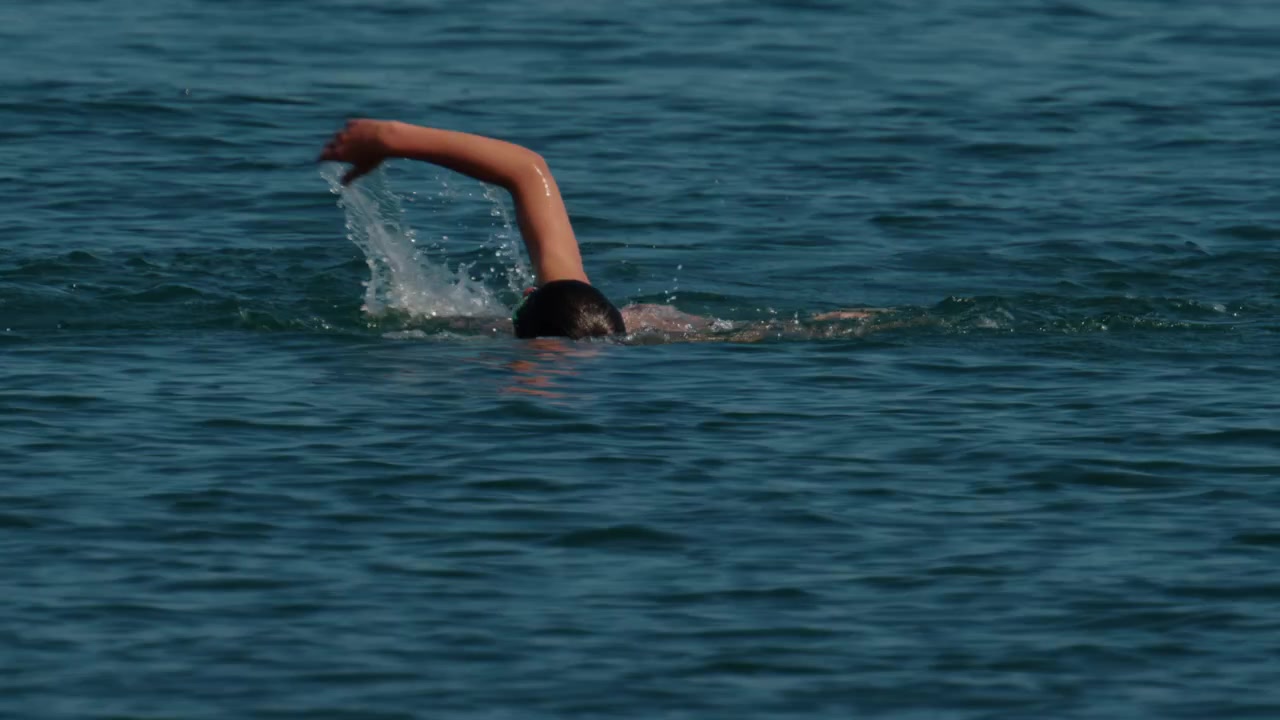 Persona nadando estilo libre en el mar, creando salpicaduras. Representa entrenamiento, resistencia, libertad y un estilo de vida activo.