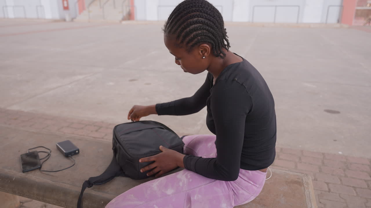 Female sorting items outside. Woman tidying backpack contents on city sidewalk during day. Young lady methodically organizing cables and phone on pavement before leaving for class or commute