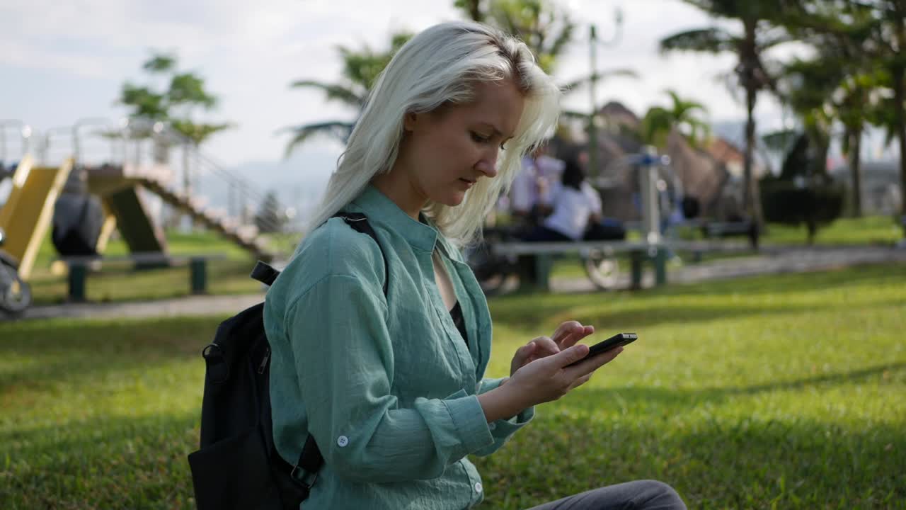 Beautiful slim woman with long blonde hair in green shirt sits on the ground and using smartphone over background the park. Girl on the square touching screen and smile