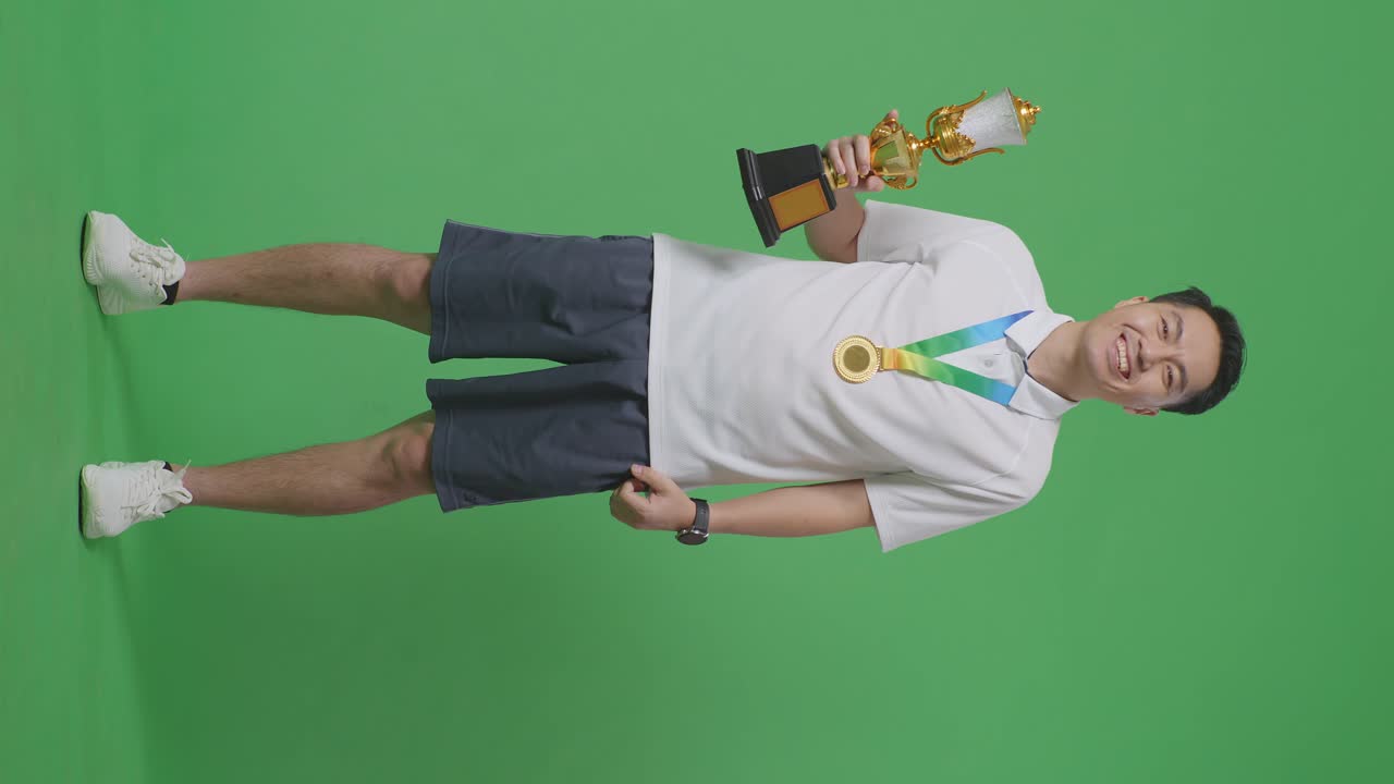 아시아 남성이 금메달을 입고 손에 금 트로피를 들고 첫 번째 우승자로 스튜디오의 녹색 화면 배경에서 카메라에 미소 짓고 있습니다.