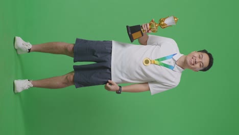 아시아 남성이 금메달을 입고 손에 금 트로피를 들고 첫 번째 우승자로 스튜디오의 녹색 화면 배경에서 카메라에 미소 짓고 있습니다.