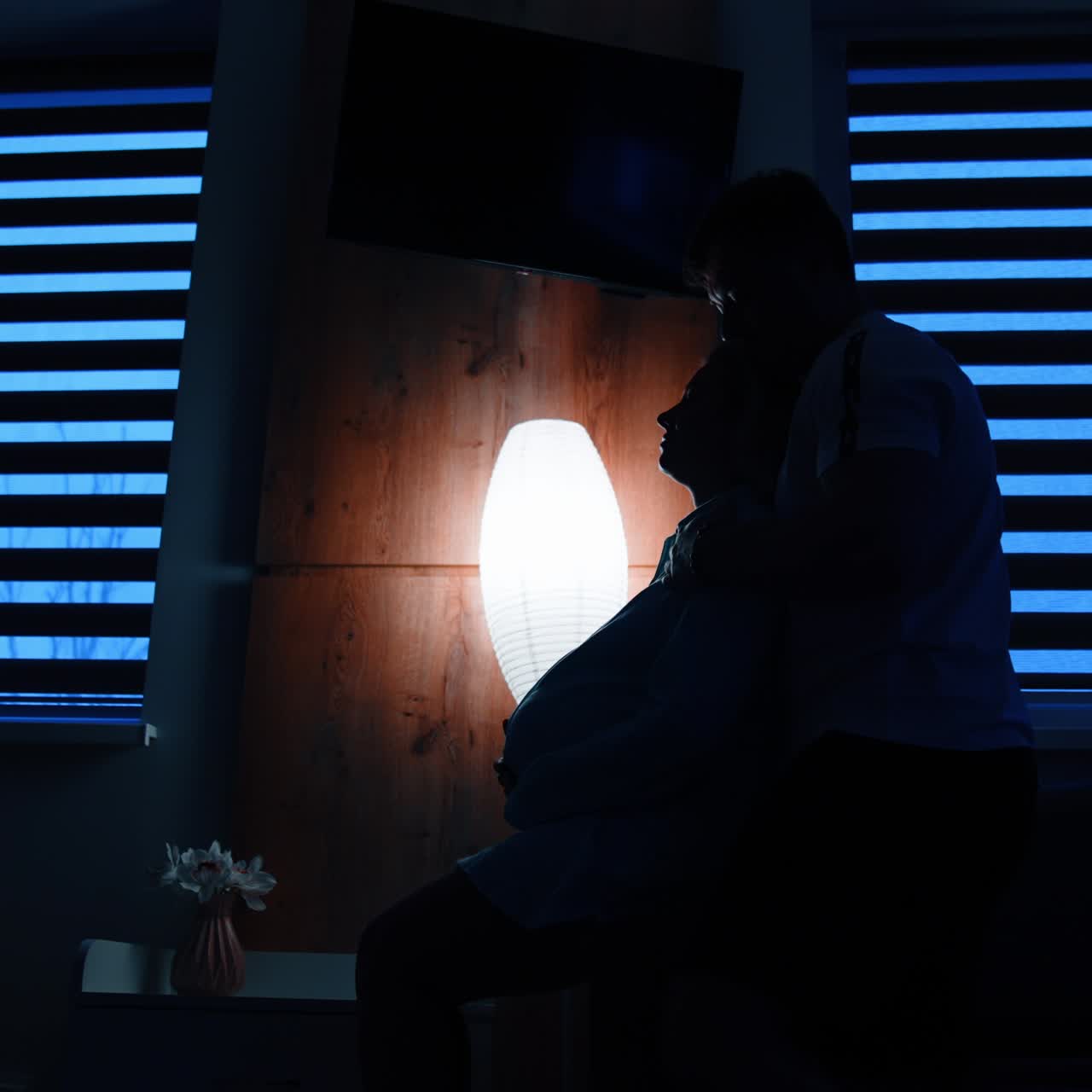 Pregnant lady sits on the bed holding by the belly. Man comes up from behind embracing a woman. Low angle view