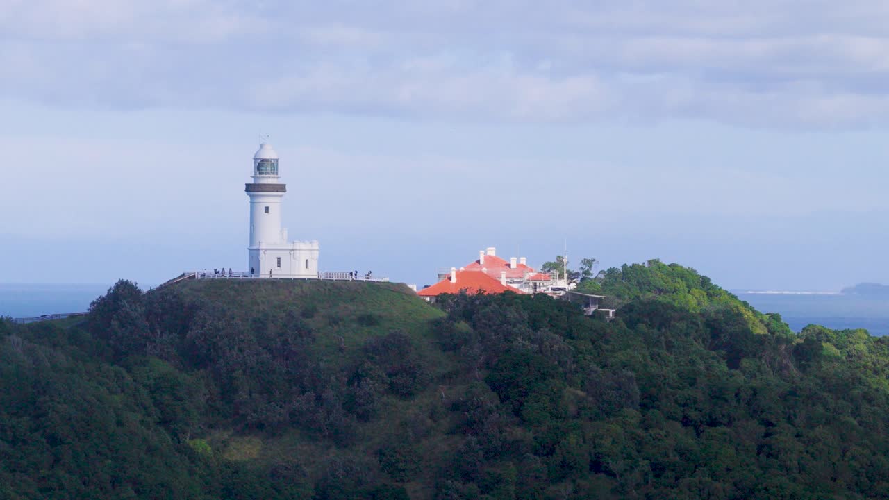 Drone footage captures Byron Bay lighthouse on a lush hilltop, surrounded by ocean, under soft evening light