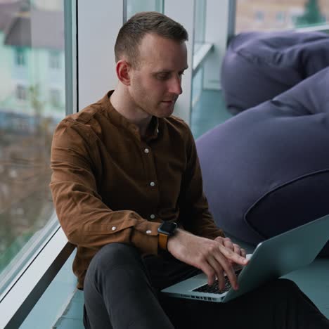 young man with laptop sitting on floor by the window. man an is working remotely
