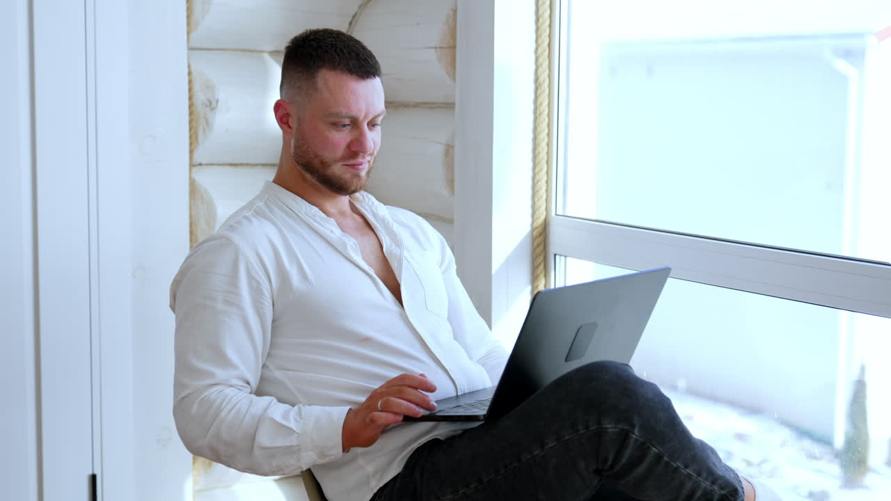 Confident relaxed Caucasian man in white shirt sits on the chair near the window. Man holds a laptop on his laps working from home.