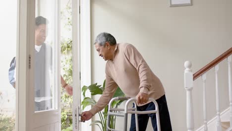 Diverse-male-doctor-and-senior-male-patient-using-walking-frame-shaking-hands-at-home,-slow-motion