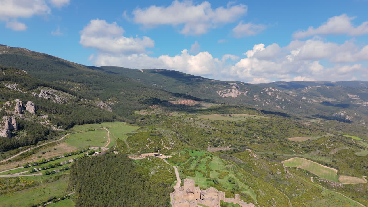 Reversing aerial shot revealing a historic stone castle on a rocky outcrop, surrounded by mountains, valleys, forest and fields under a vibrant blue sky