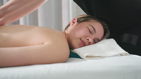 Massage therapist using firm technique to massage client back while client rests head on soft white towel in peaceful spa room with gentle light, expressing calm and relaxation during therapy session
