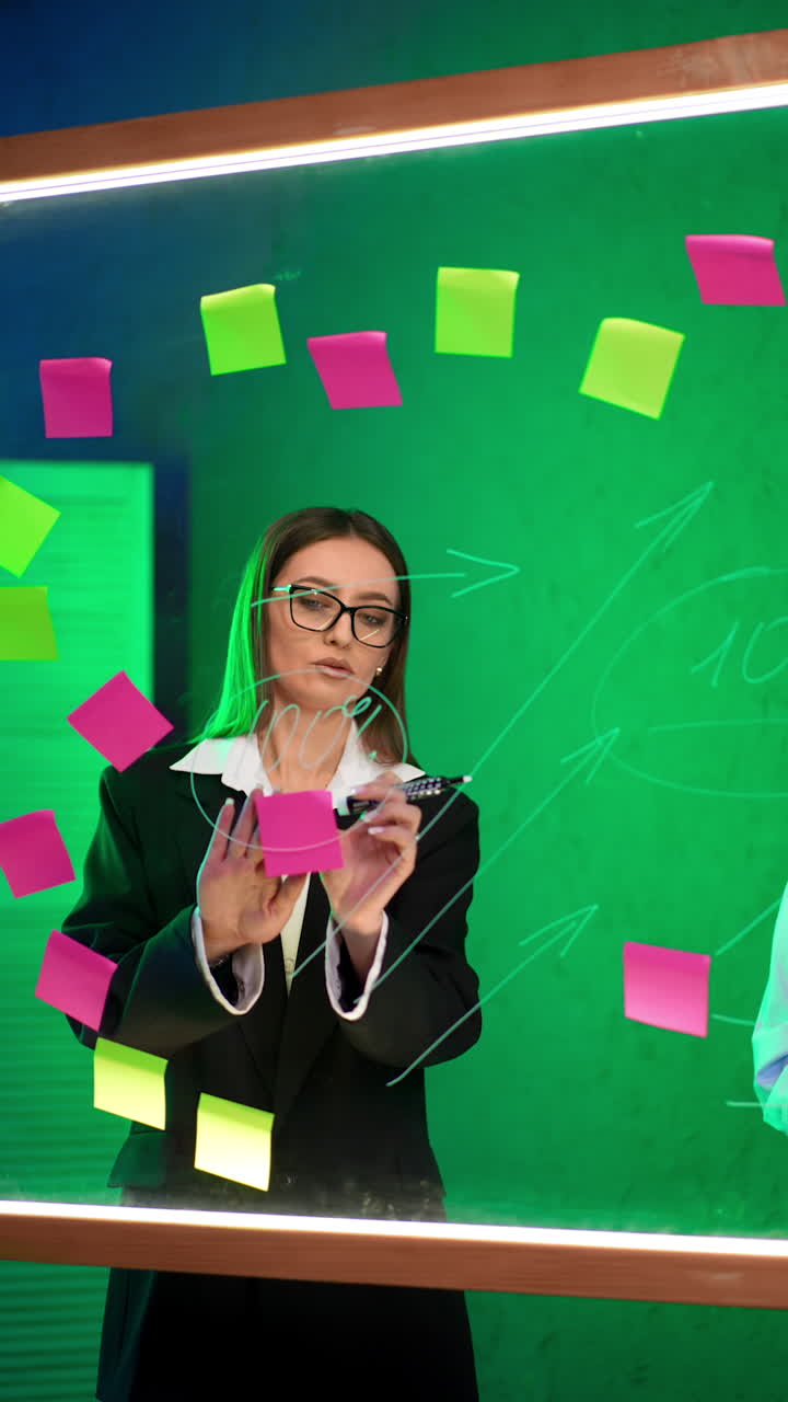 Two ladies consult at the glass board in the office. Women discuss numbers and ideas written on the stickers. Vertical video.