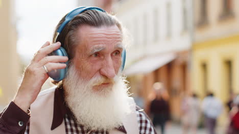 hombre mayor feliz con auriculares inalámbricos eligiendo, escuchando música bailando al aire libre en la calle de la ciudad