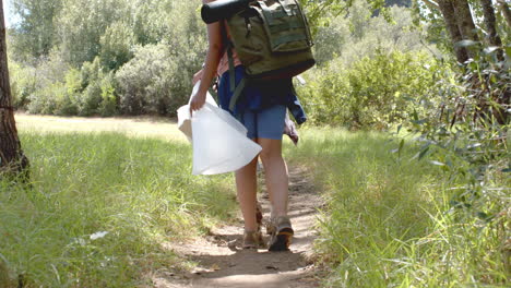 una persona está caminando por un sendero de tierra llevando una mochila grande y un contenedor blanco, recolectando basura