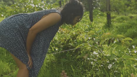 woman with black hair, wearing blue dress, bending fowards to smell aroma of plants