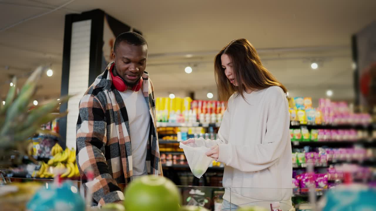una chica morena en un suéter blanco junto con su novio con piel negra en una camisa a cuadros y auriculares inalámbricos rojos eligen las limas adecuadas en un gran mostrador con frutas cítricas en un gran supermercado moderno