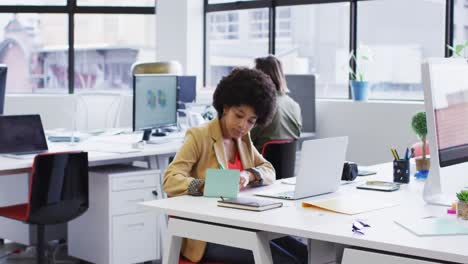 Mixed-race-businesswoman-sitting-using-a-laptop-going-through-paperwork-in-modern-office