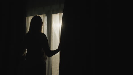 Silhouette-of-a-woman-who-opens-blackout-curtains.-It-is-illuminated-by-the-bright-sun-from-the-window.-Back-view